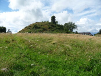 In a grassy field, the earthwork remains of Ewyas Harold Castle, rises against a blue cloudy sky.